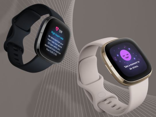Dois smartwatches Fitbit, um preto e outro marfim com caixa dourada, flutuando sobre um fundo cinza-quente